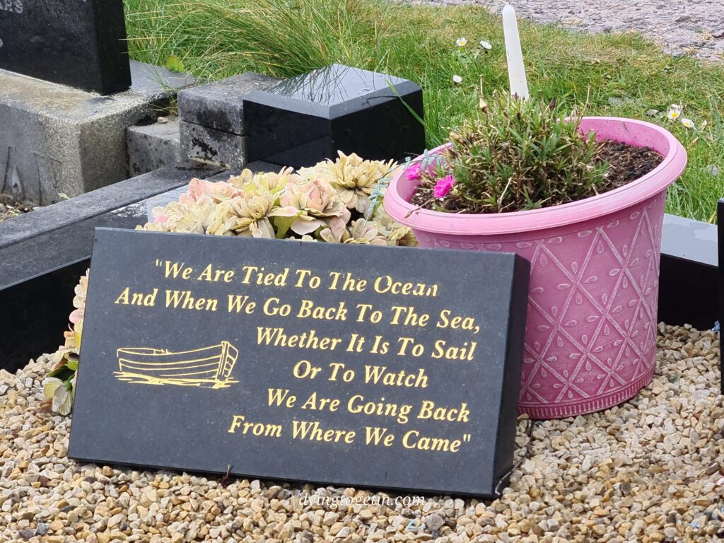 Black plaque on which is written in gold: We are tied to the ocean. And when we go back to the sea, whether it is to sail or to watch - we are going back from whence we came. It's set on pebbles on a grave proppsed against a pink flowerpot from which sticks a long white candle