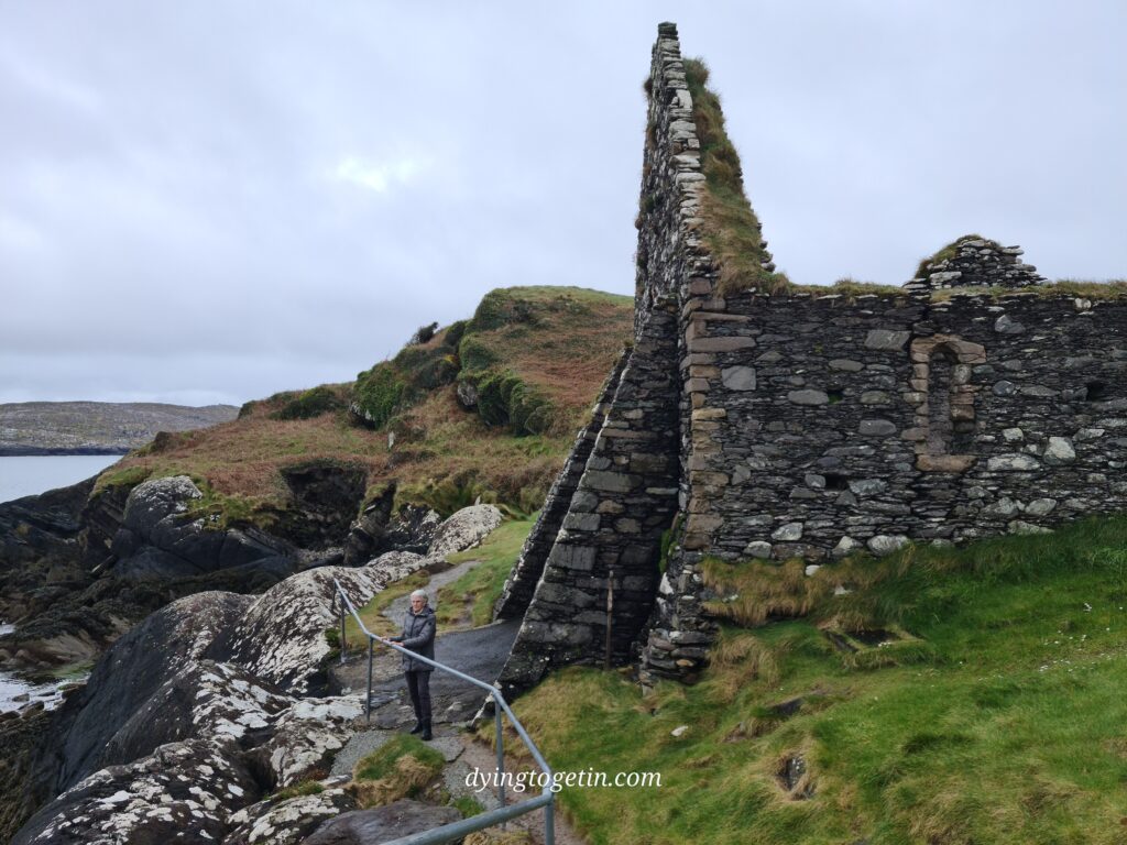 White haired woman stands facing the sea, her back to the ruins of an old church