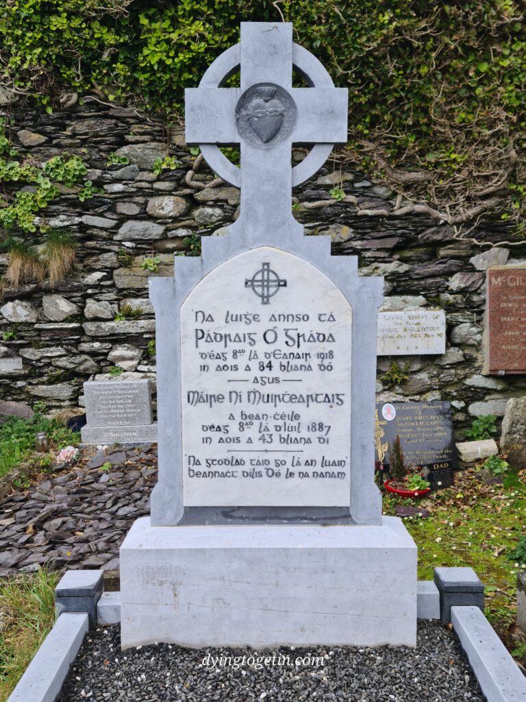 Granite cross headstone with a heart in the middle. Inscription is in Irish and reads: N Luige annso tá Padraig O'Gráda deag 8 la b'eanair 1918 in aois a 84 bliana dó. Máire ni Muinteartaig a bean-céile, déag 8 lá d;Iuil 1887 in aois a 43 Bliana dí Na gcodlad táid go lá an luain beannache dilis dé la na nanam. Other headstones visible in the background against a stone wall
