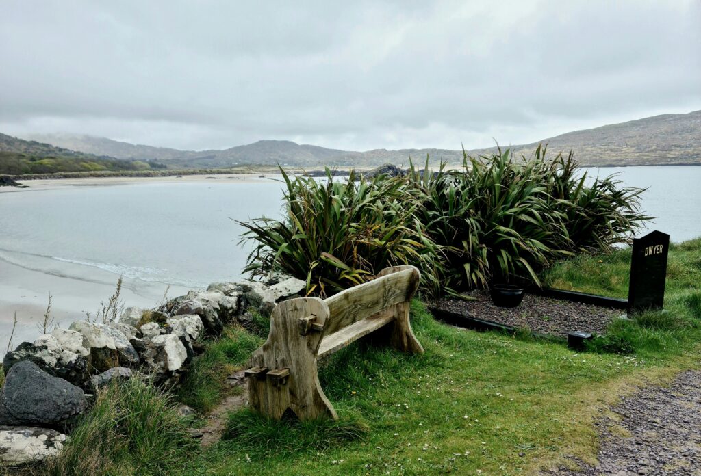 Wooden bench on a grassy knoll behind a stone wall overlooking the sea. Next to it is a black marble headstone with the word DWYER etched in gold. 