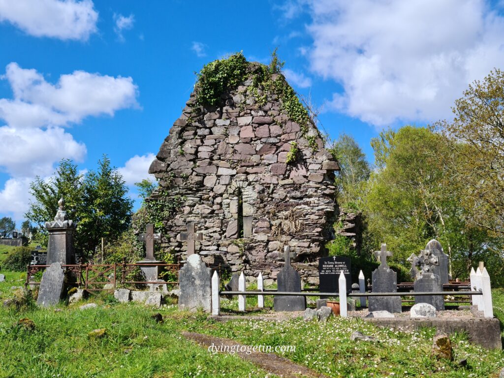 The gable wall of an old stone church surrounded by headstones. A blue cloudy sky tops the photo and an expanse of green grass tails it.