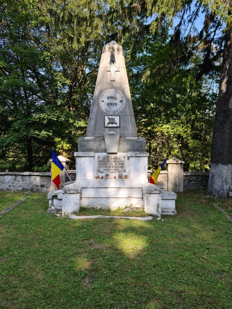Stone monument with a cross over a circle with the dates 1916 1919. Under the circle is a soldiers helmet and crossed swords. The inscription is in Romanian and reads: Aici odihnesc ostasii necunoscutu cazuti pe aceste locuri in razboiul. Two Romanian flags (blue, yellow, red) stand on either side,