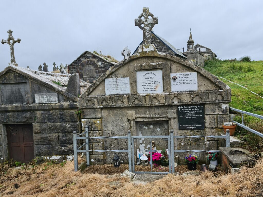 Gabled tombs one with three plaques on the wall, a grey iron fence around it, and some pink flowers in the foreground. The apex of the roof bears a celtic cross.