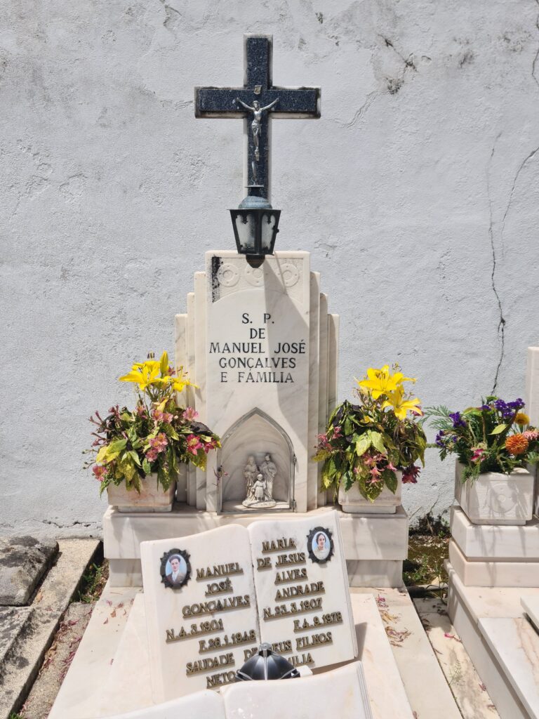 Manual Jose Goncalves and family 0 their grave. Statues the Holy family stand in a glass niche at the base. A crucifix crown the top from which hangs a lantern. Yellow flowers in posts sit either side. 