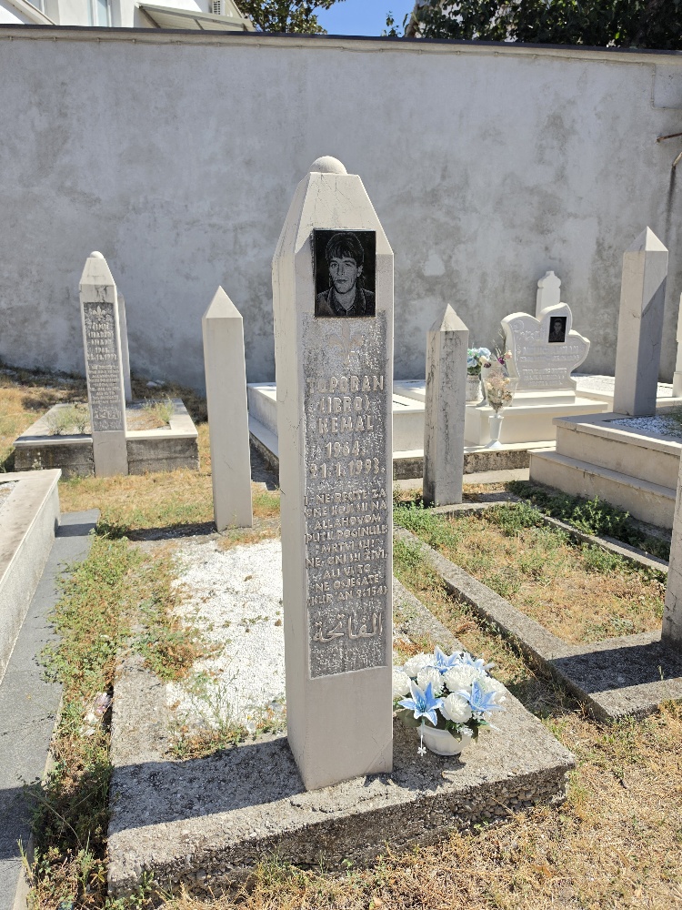 A Muslim cemetery in Mostar with white stone graves and tall, pointed headstones. In the foreground is the grave of Kemal Toporan (1964–21 January 1993), with his photograph on the headstone. Other graves are visible behind him.