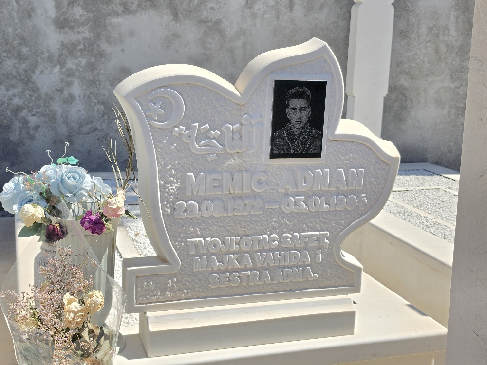 Close-up of a white, ornate Muslim gravestone bearing a photograph of a young man. The inscription reads “Memlić Adnan, 29.08.1972 – 03.01.1993”, followed by a message from his father, mother, and sister. A crescent-and-star symbol appears near the top.