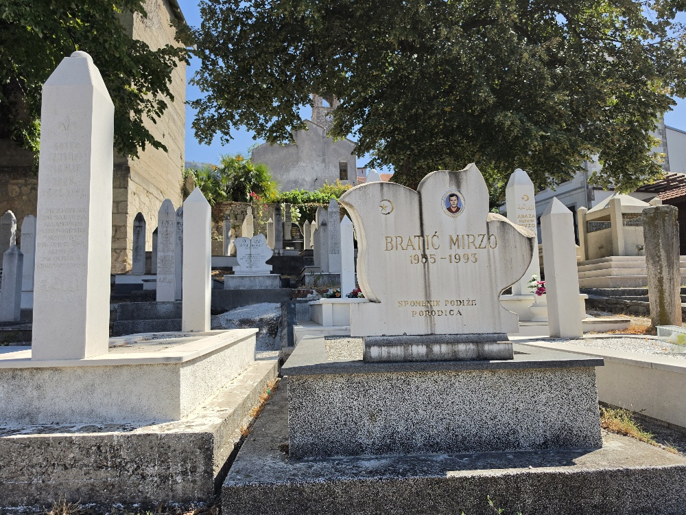 “A white marble grave monument for Mirzo Bratić (1955–1993), with a curved decorative design, crescent-and-star symbol, and portrait photograph, surrounded by tall upright Muslim gravestones.”