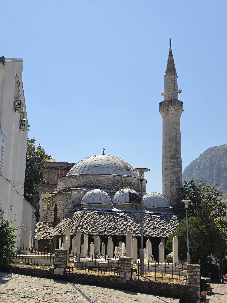 “White Muslim gravestones in Šehitluci cemetery in Mostar, with a historic mosque featuring several domes and a tall stone minaret behind them, and a steep mountain visible to the right.”