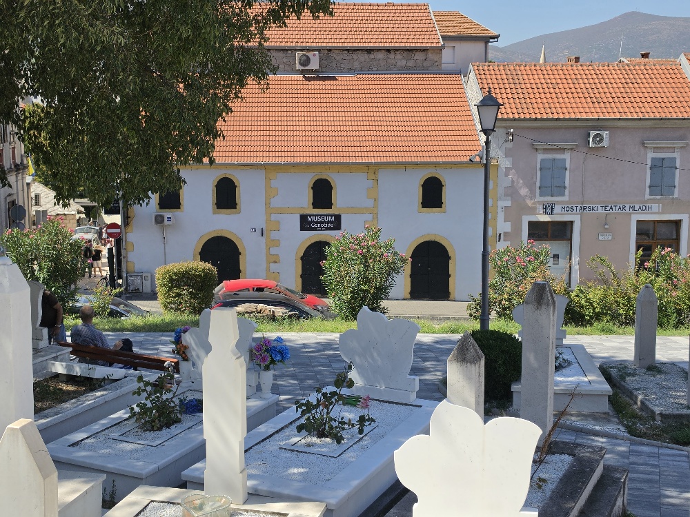 “Graves in Šehitluci cemetery overlooking a street in Mostar, with white Muslim gravestones in the foreground and the Museum of War and Genocide Victims 1992–1995 in a historic building across the street.”