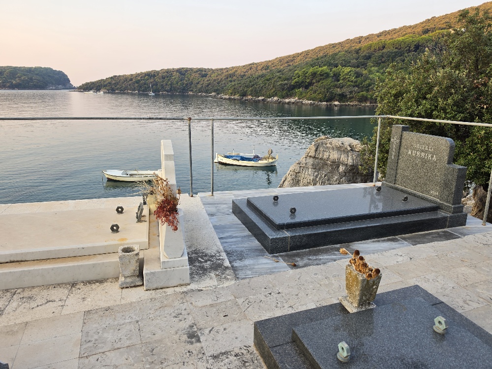 Graves in Molunat Cemetery overlooking a calm Adriatic bay, with small boats on the water and wooded hills on the opposite shore.