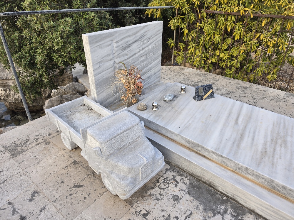 A white marble grave in Molunat Cemetery decorated with flowers, small stones and a truck beside the tomb.