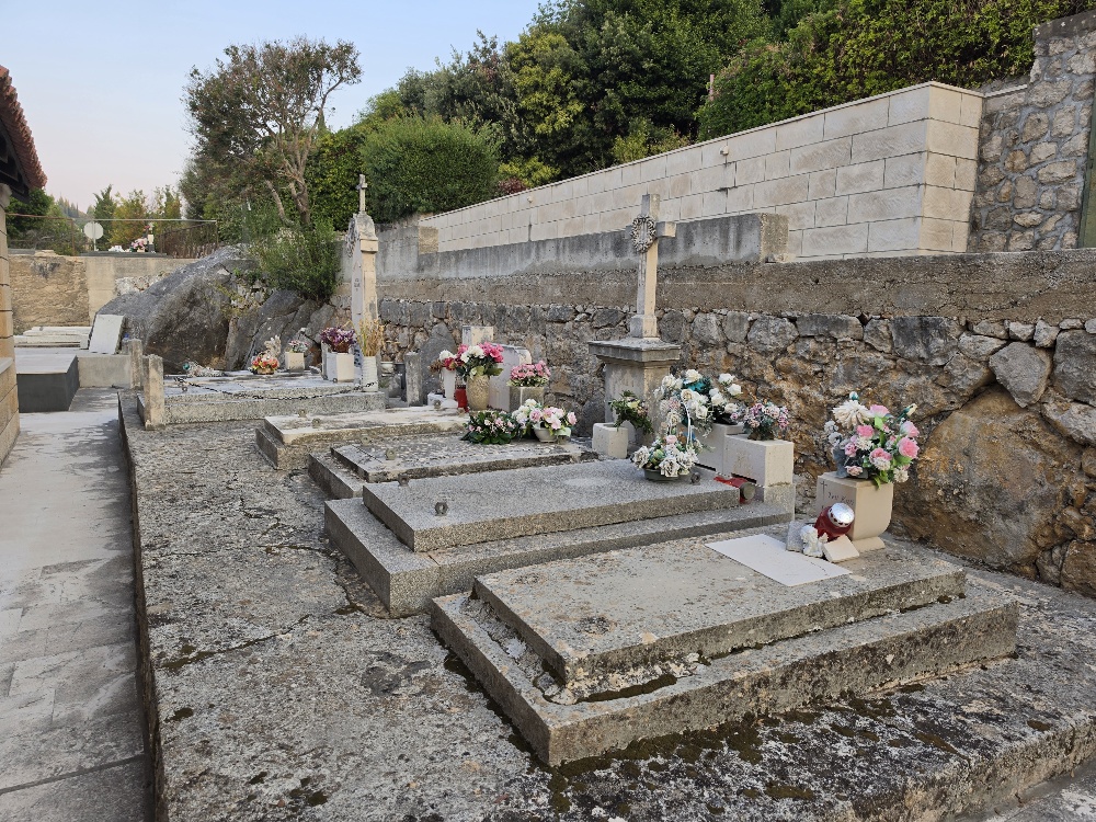 A view across Molunat Cemetery showing rows of stone family tombs beside the small coastal church, with the Adriatic bay and wooded shoreline beyond.