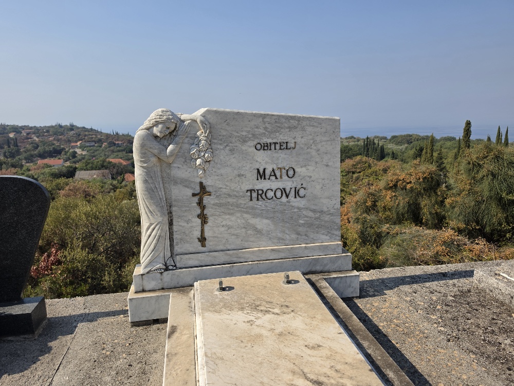 Marble family tomb marked “Obitelj Mato Trcović” in the cemetery at Vitaljina, decorated with a carved mourning figure, floral ornament and crucifix, with the Konavle hills and Adriatic Sea in the background.